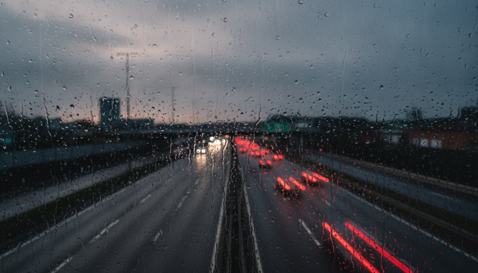 A rain-streaked window overlooking a grey motorway at dusk with blurred red tail-lights